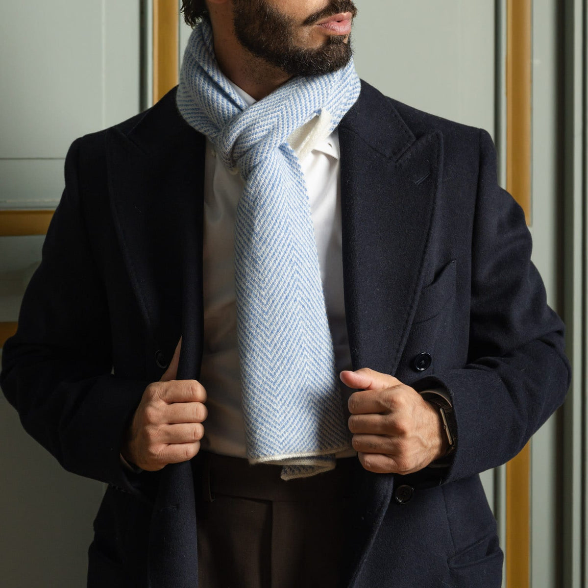 A man wearing a navy wool blazer, white dress shirt, and a light blue and white herringbone patterned scarf, standing indoors.