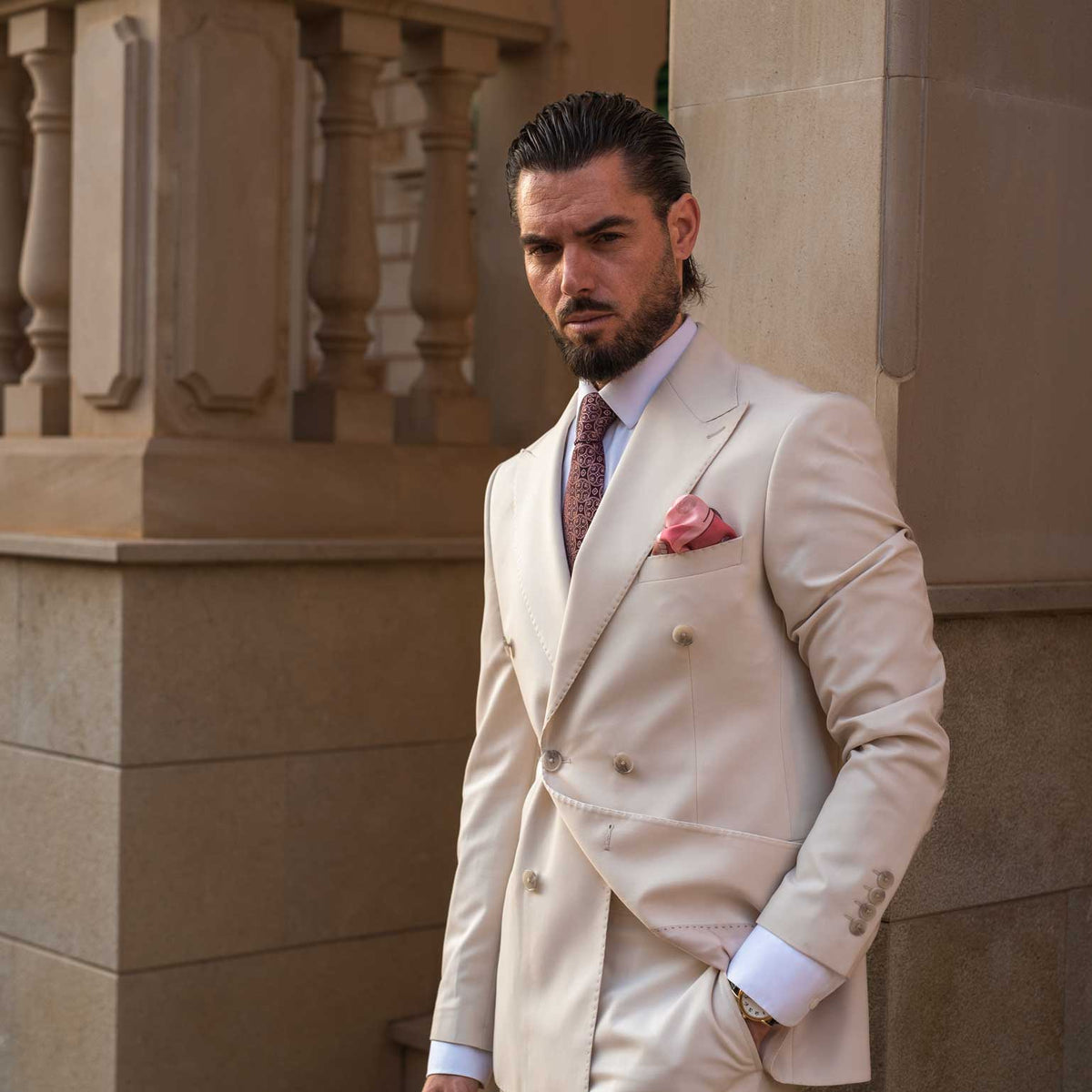 Man wearing a burgundy silk tie with a pink pocket square standing against a stone wall.
