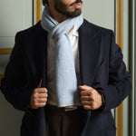 A man wearing a navy wool blazer, white dress shirt, and a light blue and white herringbone patterned scarf, standing indoors.